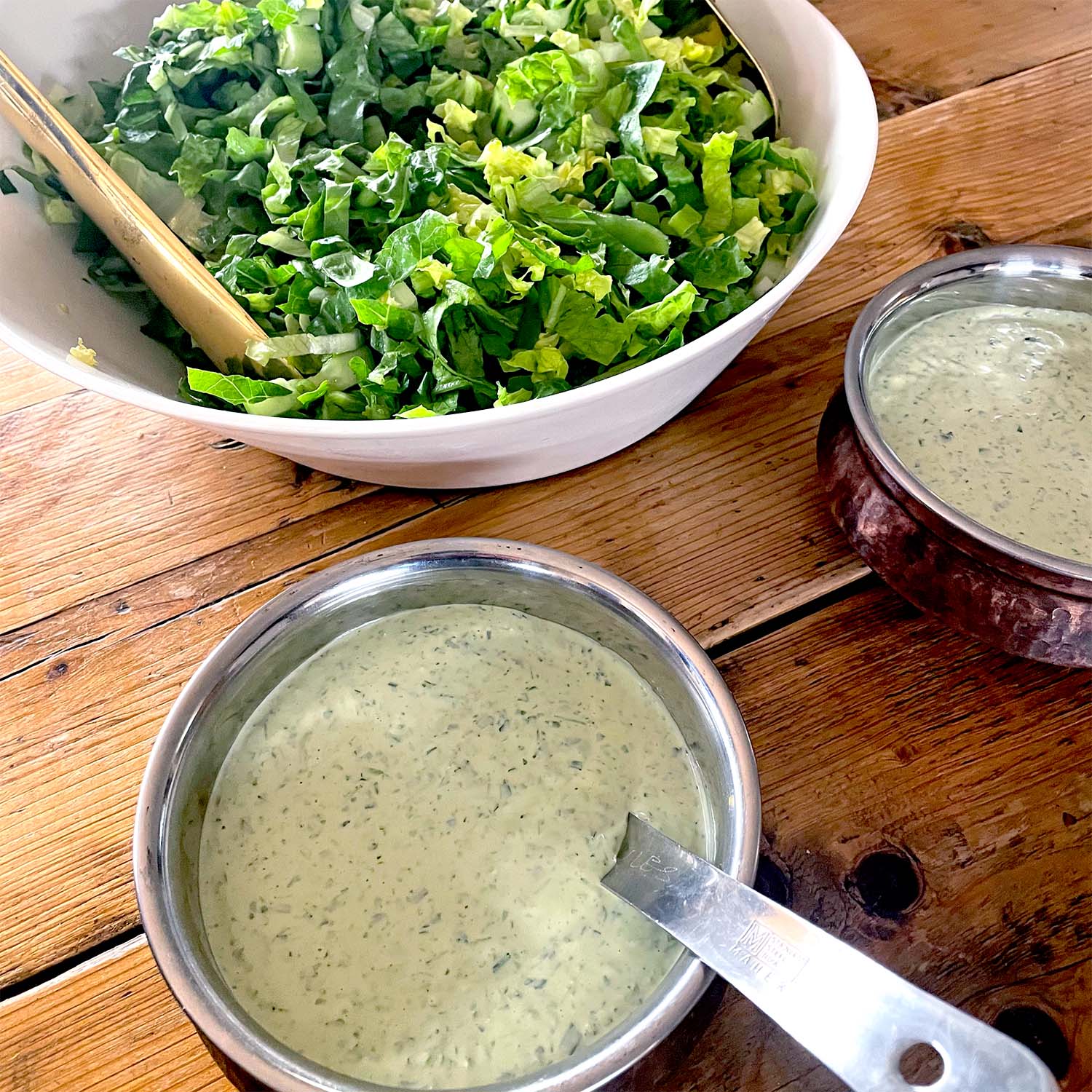 A bowl of chopped green salad with tongs next to two bowls of creamy green herb dressing on a wooden table.