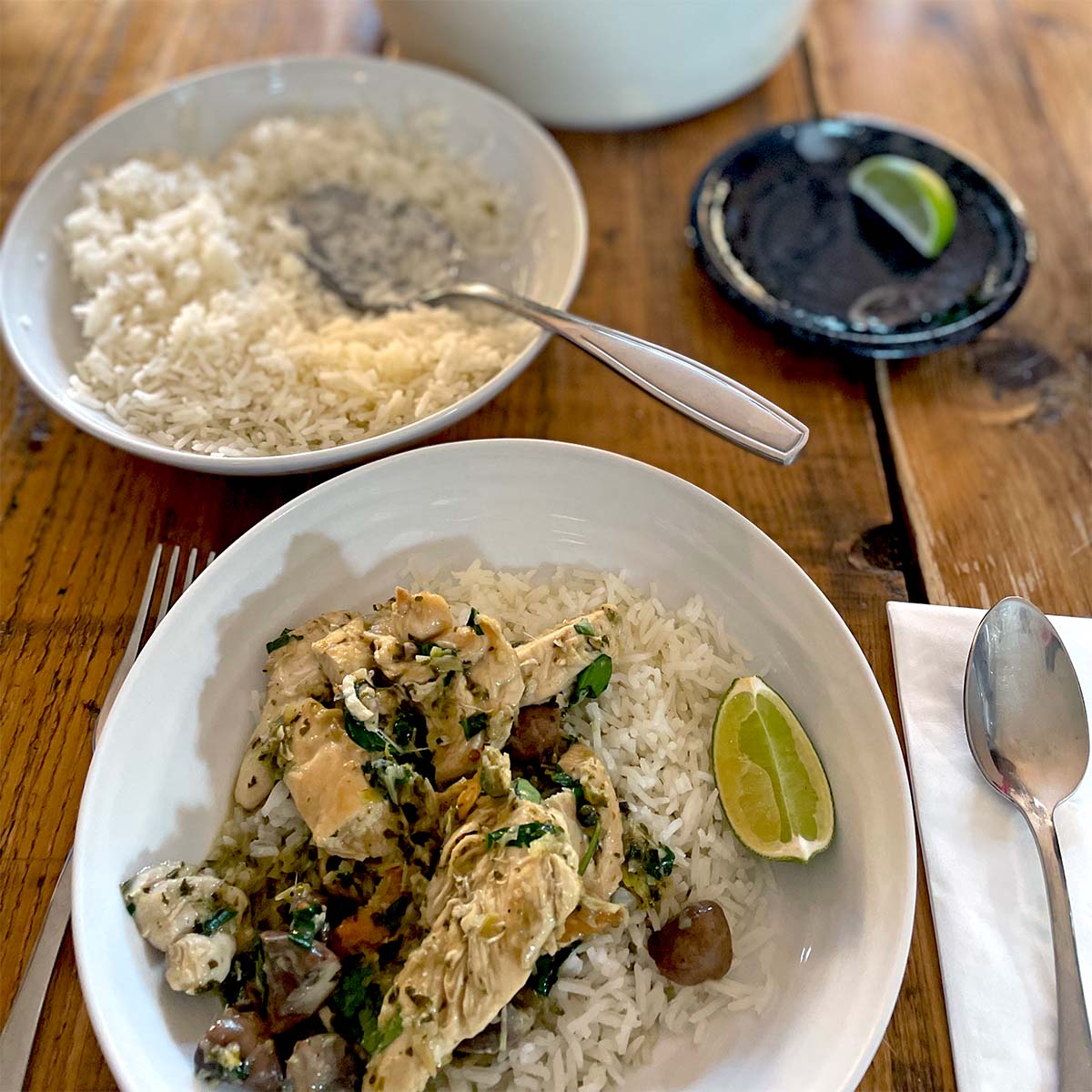 A bowl of white rice topped with chicken, vegetables, and herbs sits on a wooden table next to a lime wedge, spoon, serviette, plate with another lime wedge, and a bowl of plain rice with a spoon.