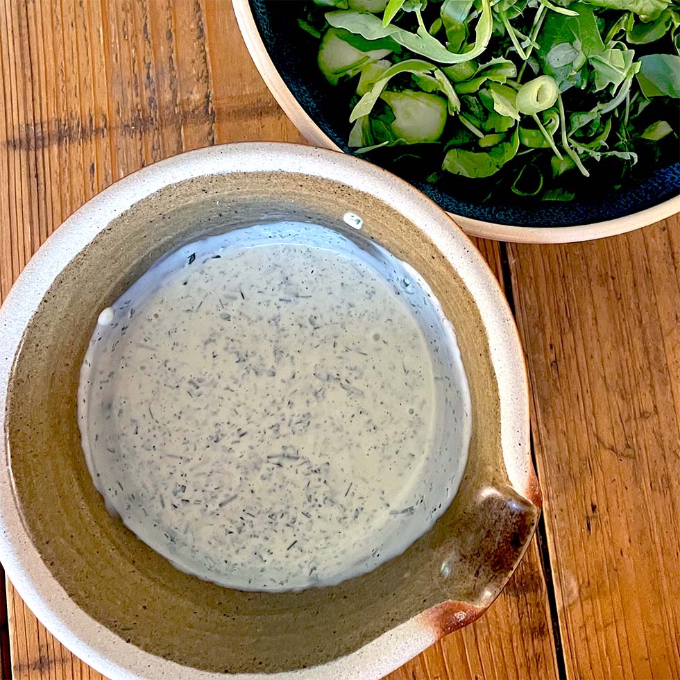 A rustic ceramic bowl filled with creamy herb dressing sits on a wooden table next to a dark bowl of fresh rocket and cucumber salad.