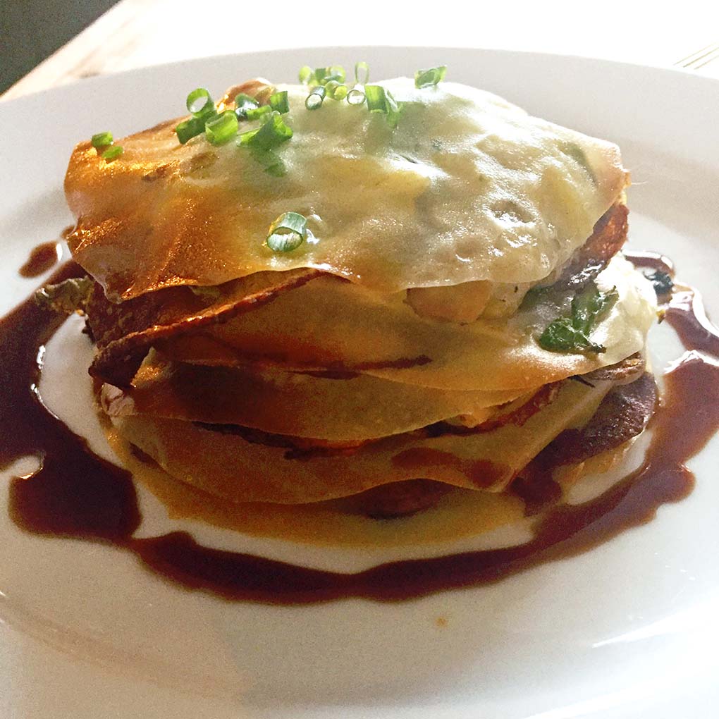 A plated dish featuring a layered savoury pastry with a golden-brown top, garnished with chopped spring onions and surrounded by a dark sauce on a white plate.