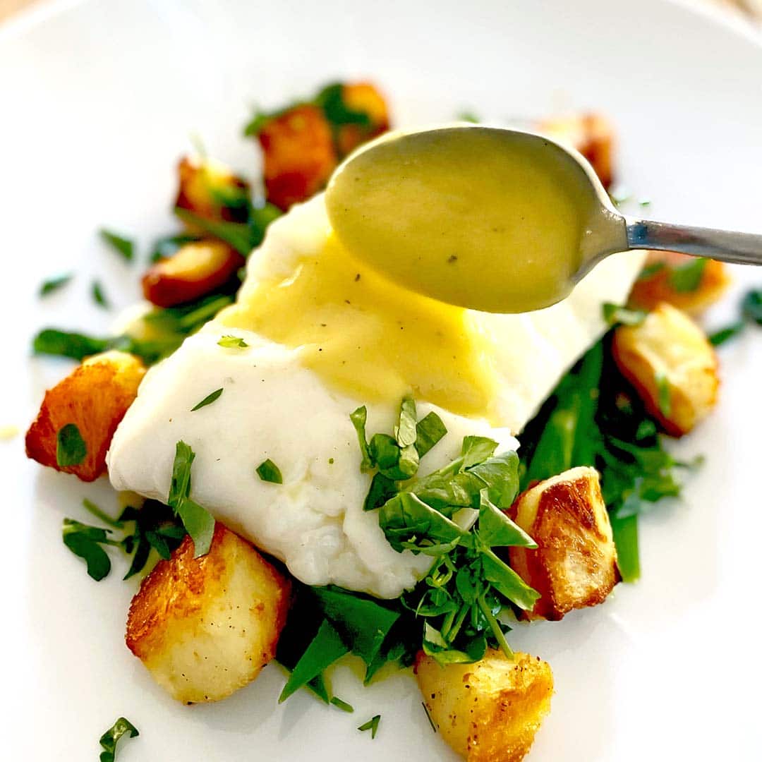A spoon drizzles sauce over a piece of white fish on a bed of green beans and roast potatoes, garnished with fresh chopped herbs, served on a white plate.