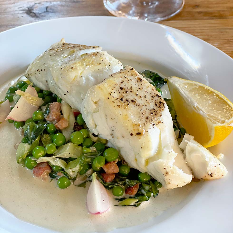 A white plate with two pieces of seasoned, cooked white fish on a bed of green peas, leafy greens, and diced vegetables, garnished with a lemon wedge and served with a light sauce.