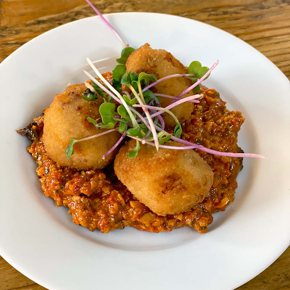 Three golden brown, breadcrumbed croquettes served on a bed of chunky red sauce, garnished with fresh green microgreens, on a white plate set on a wooden table.