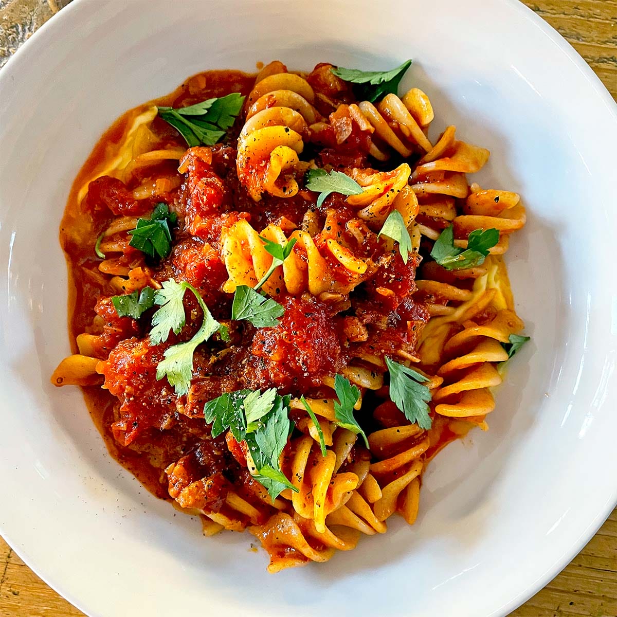 A white bowl filled with fusilli pasta topped with chunky tomato sauce and garnished with fresh parsley leaves, placed on a wooden surface.