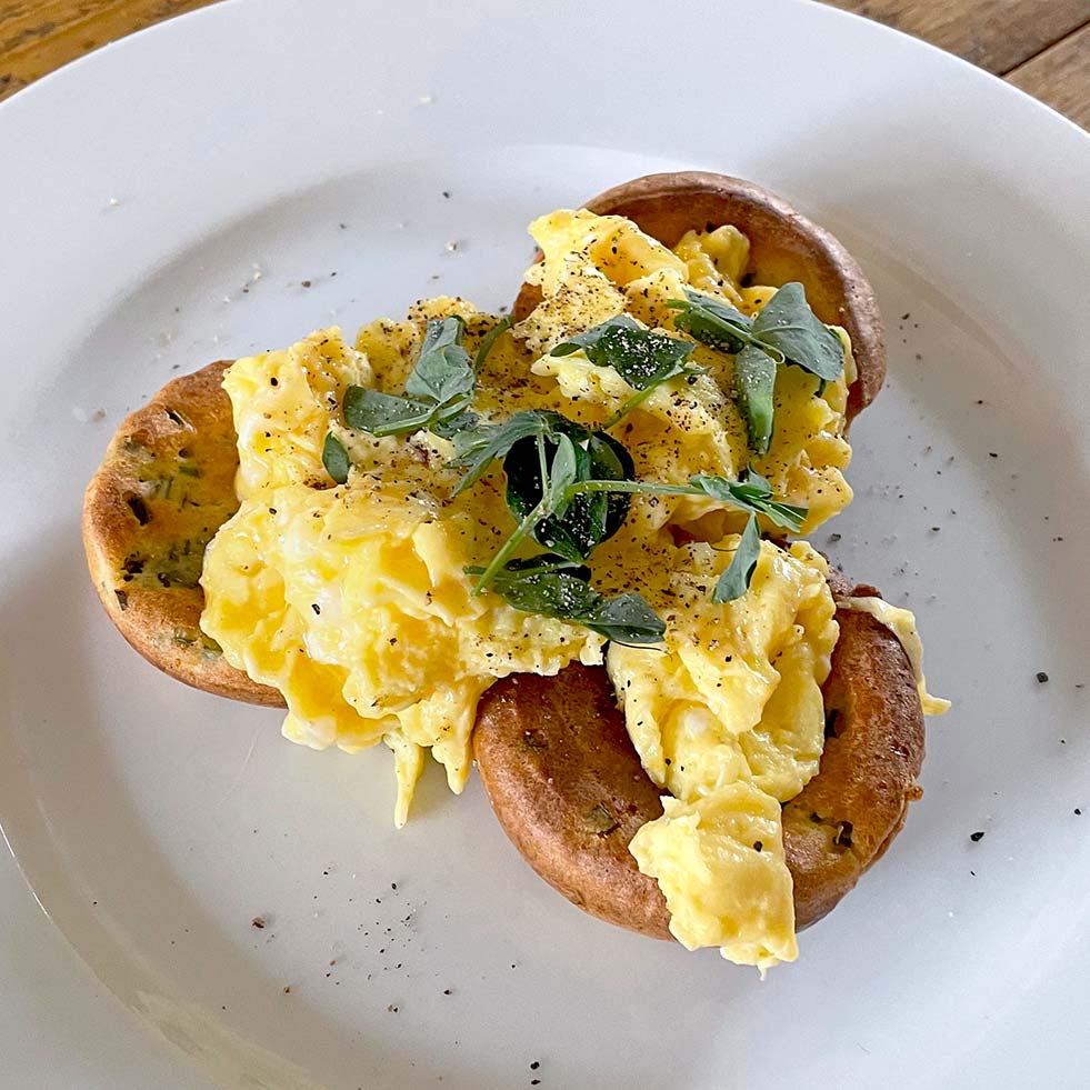 Scrambled eggs served on two toasted crumpets, garnished with fresh herbs and cracked black pepper, on a white plate.