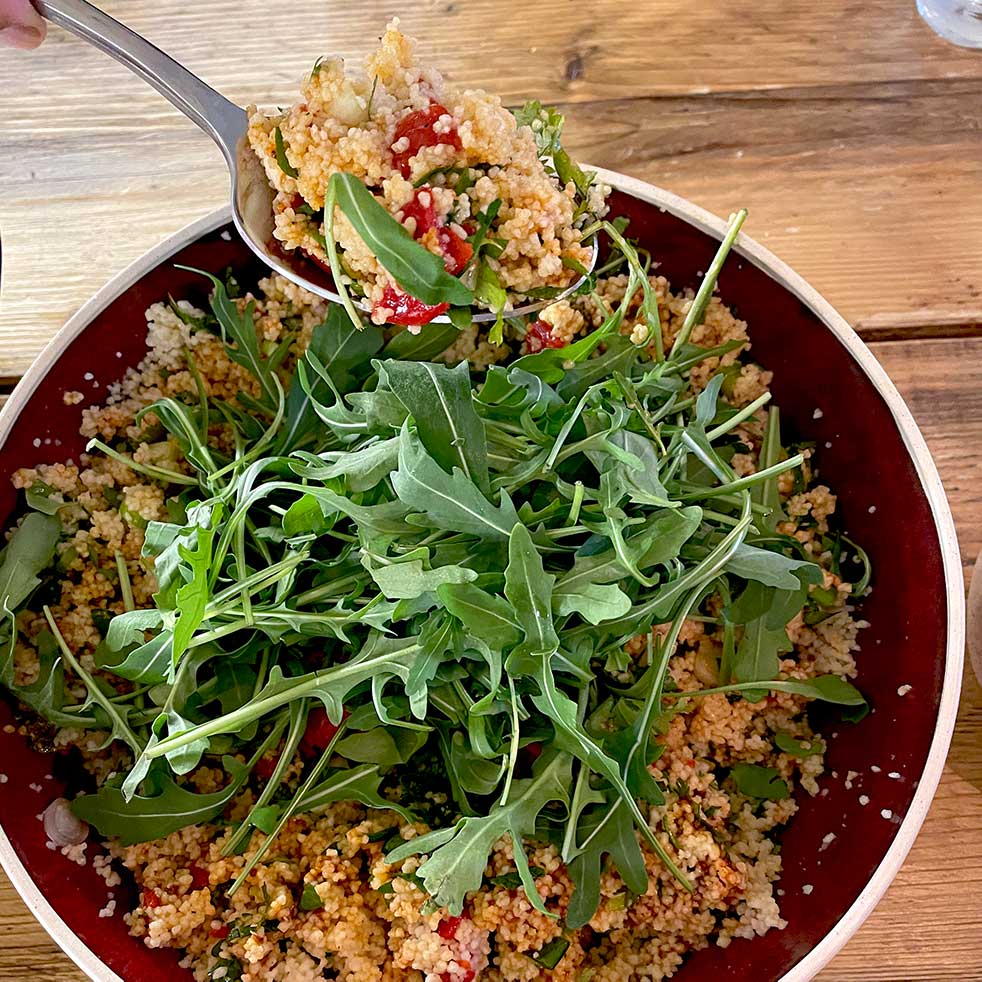 A bowl of couscous salad with chopped vegetables, garnished with fresh rocket leaves, sits on a wooden table. A spoon is serving a portion of the salad from the bowl.
