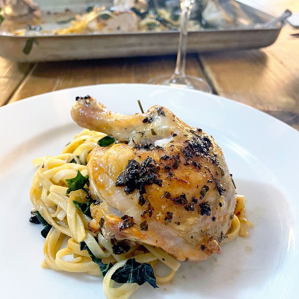 A roasted chicken leg, seasoned with herbs, is served on a bed of fettuccine pasta with greens on a white plate. In the background, a tray with more chicken and a glass of wine can be seen.