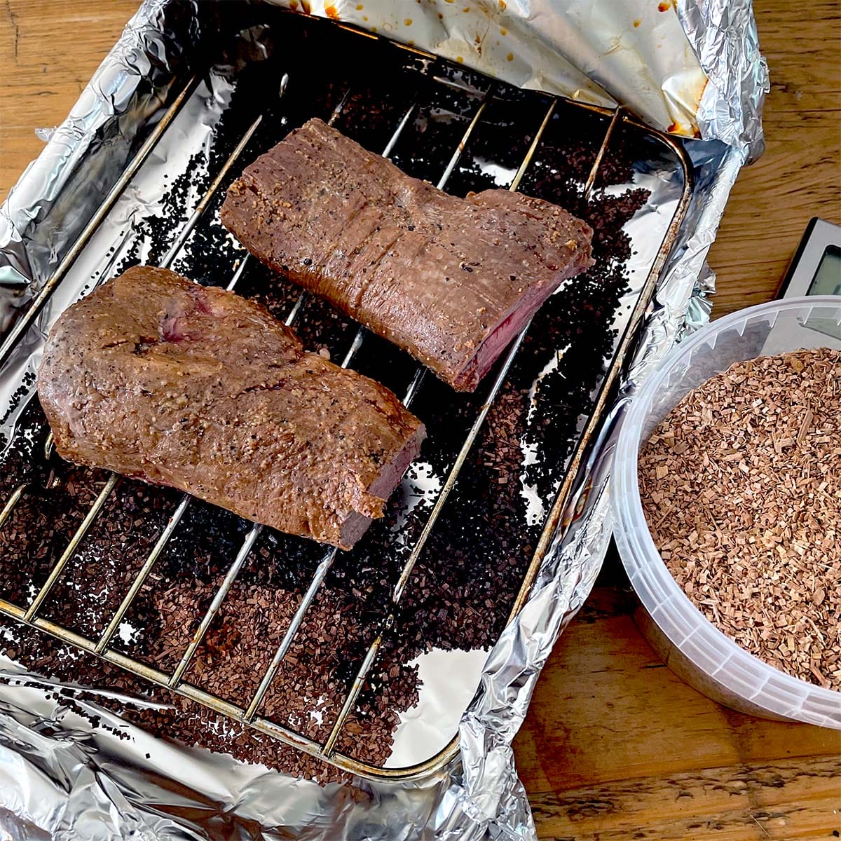 Two pieces of cooked meat rest on a metal rack inside a foil-lined roasting tin. Beside the tin is a plastic container filled with wood chips, likely for smoking the meat.