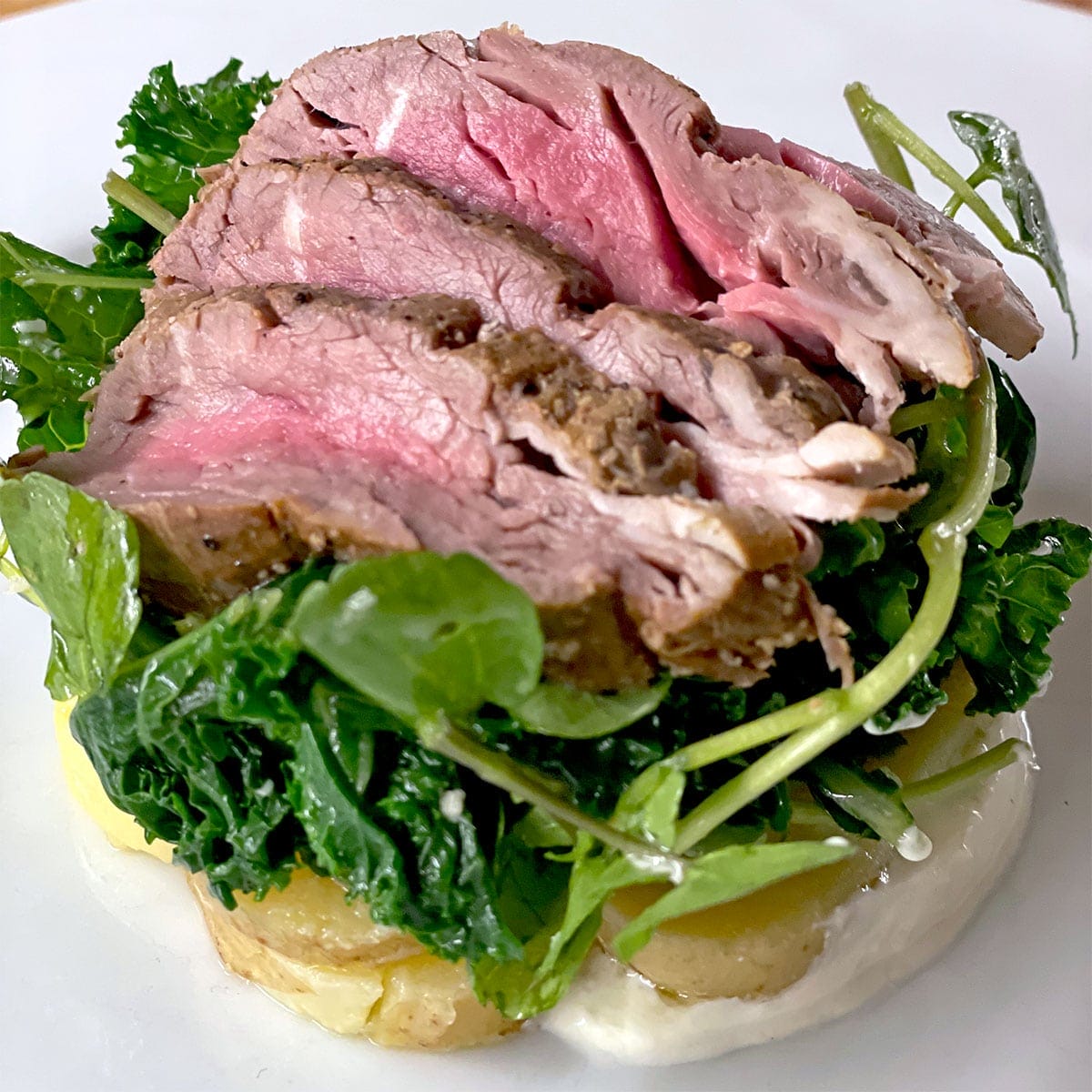 Slices of medium-rare roast beef are served on a bed of leafy greens and watercress atop sliced potatoes with a creamy white sauce, arranged on a white plate.
