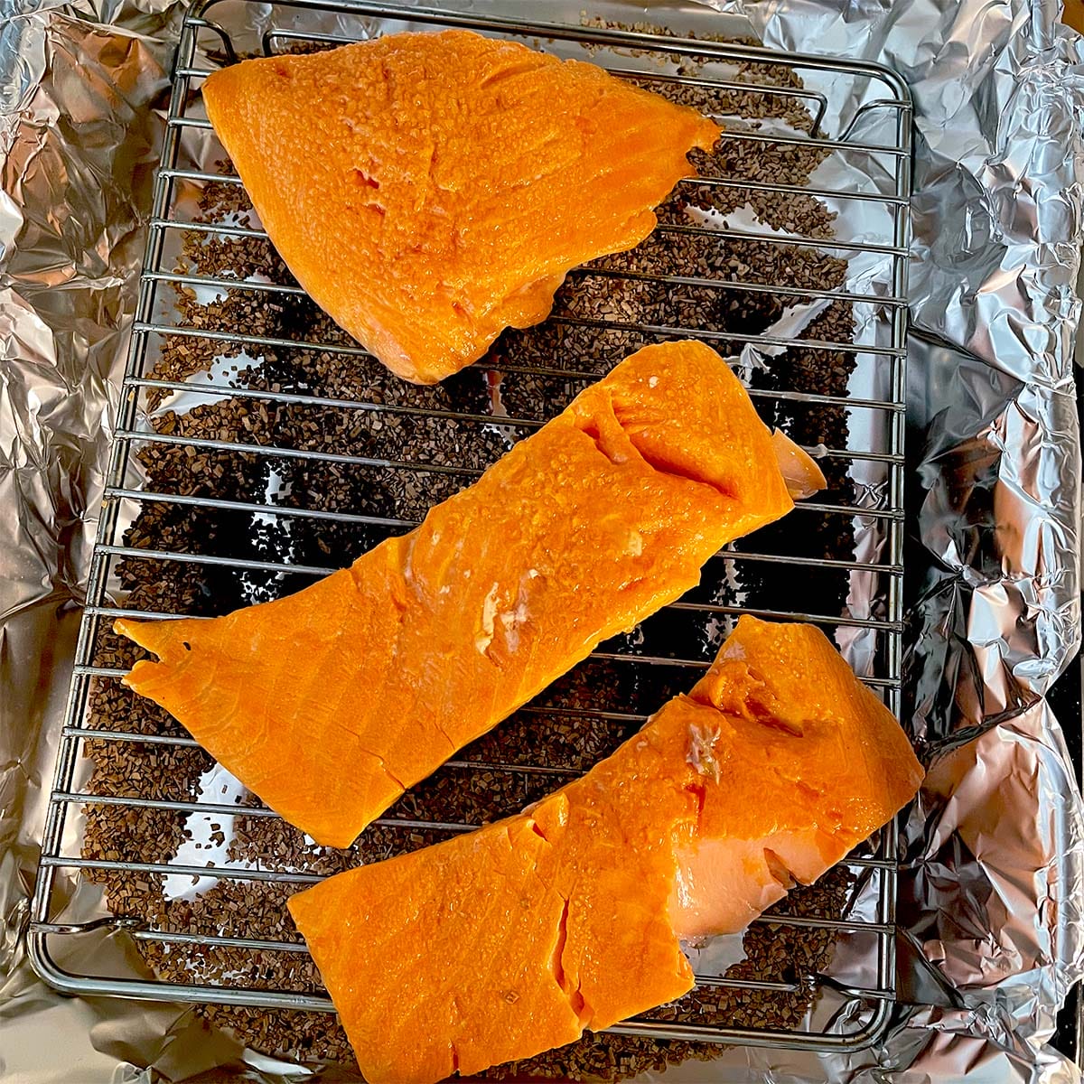 Three pieces of raw salmon fillet are placed on a metal rack over a layer of brown wood chips on aluminium foil, ready to be smoked or cooked.