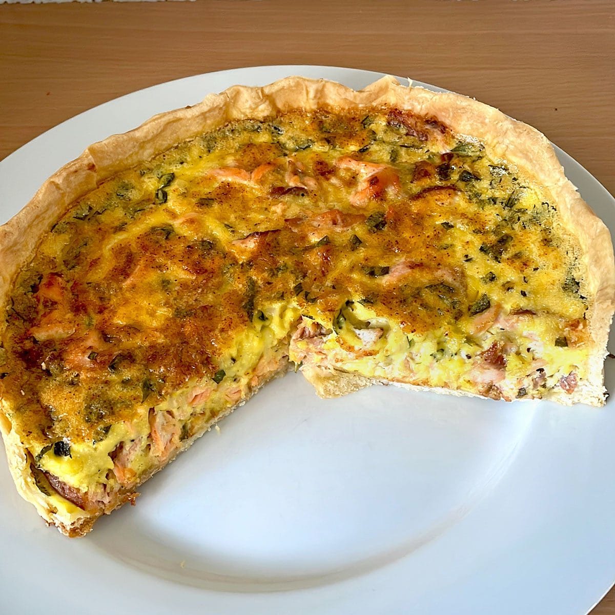 A baked quiche with a golden crust and a savoury filling, featuring visible pieces of vegetables and meat, sits on a white plate with a large slice removed.