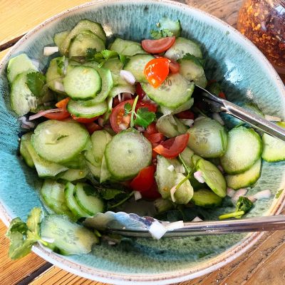 A bowl of fresh cucumber salad with sliced cucumbers, cherry tomatoes, onions, and coriander, served with metal tongs on a wooden table.