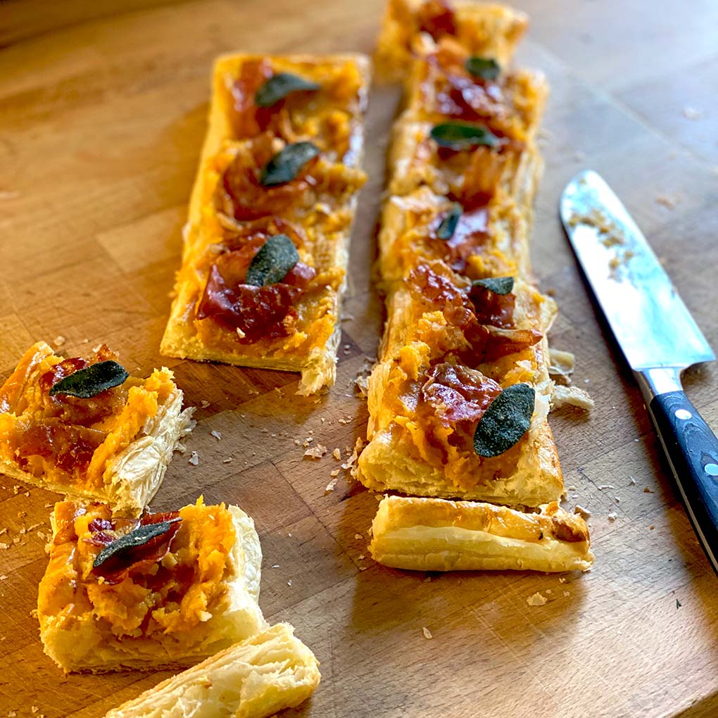 Rectangular puff pastry tart topped with orange mashed squash, crispy sage leaves, and slices of cured meat, cut into pieces on a wooden surface next to a knife.