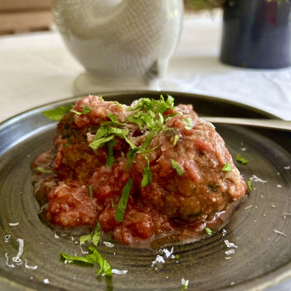 Two meatballs topped with tomato sauce and chopped fresh herbs are served on a dark plate. Some grated cheese is visible on top and around the plate, with a spoon resting beside the meatballs.