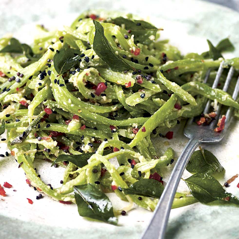 A plate of green pasta topped with black seeds, red chilli flakes, grated cheese, and fresh herbs. A fork rests on the plate beside the pasta.