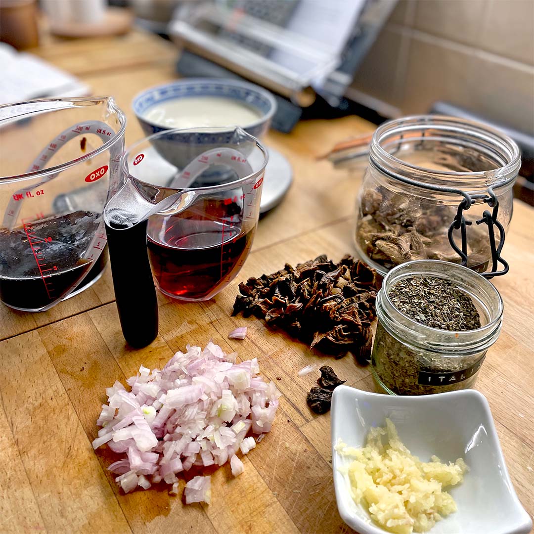 Chopped shallots, minced garlic, dried mushrooms, dried herbs, red wine, and cream are arranged on a wooden kitchen worktop, ready for cooking. Measuring jugs and glass jars hold some of the ingredients.