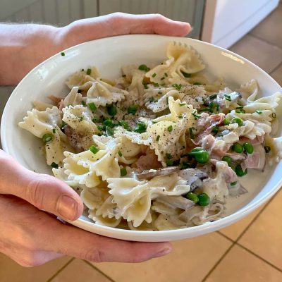 A person holds a white bowl filled with farfalle pasta in a creamy sauce, mixed with peas, ham, mushrooms, and garnished with chopped chives and ground black pepper.