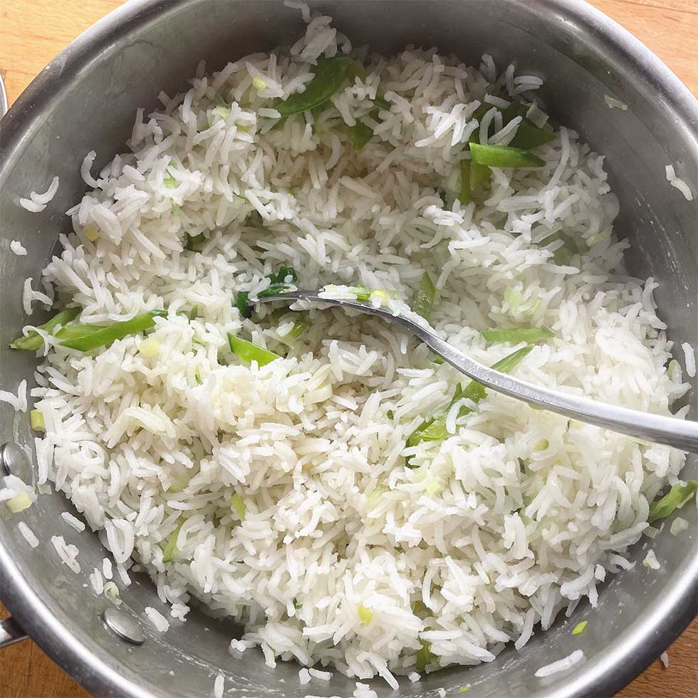 A pot of cooked white basmati rice mixed with chopped spring onions, with a metal spoon resting inside the pot.