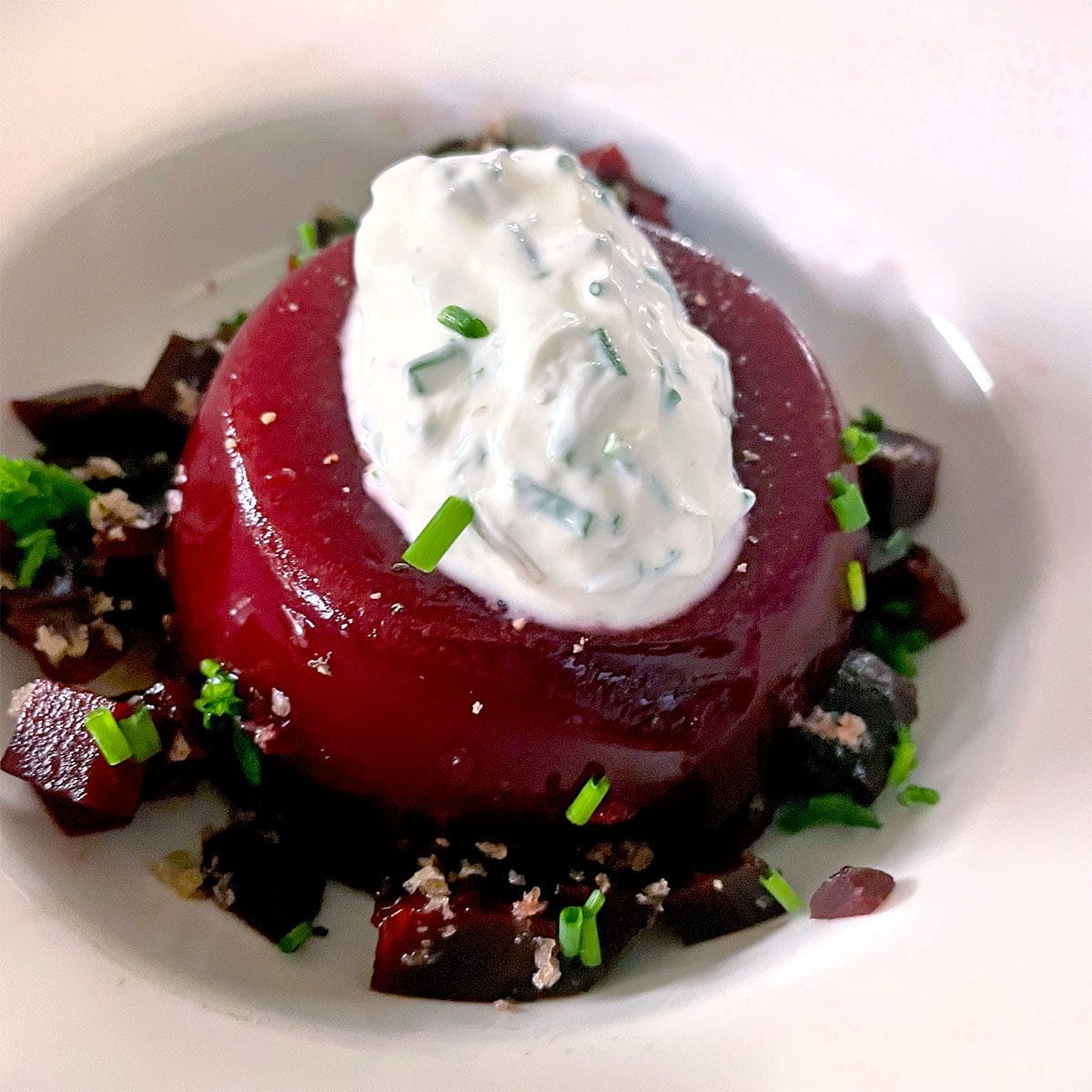 A round serving of beet jelly topped with a dollop of herbed cream sits on a plate, surrounded by small diced beetroot and sprinkled with fresh chives.