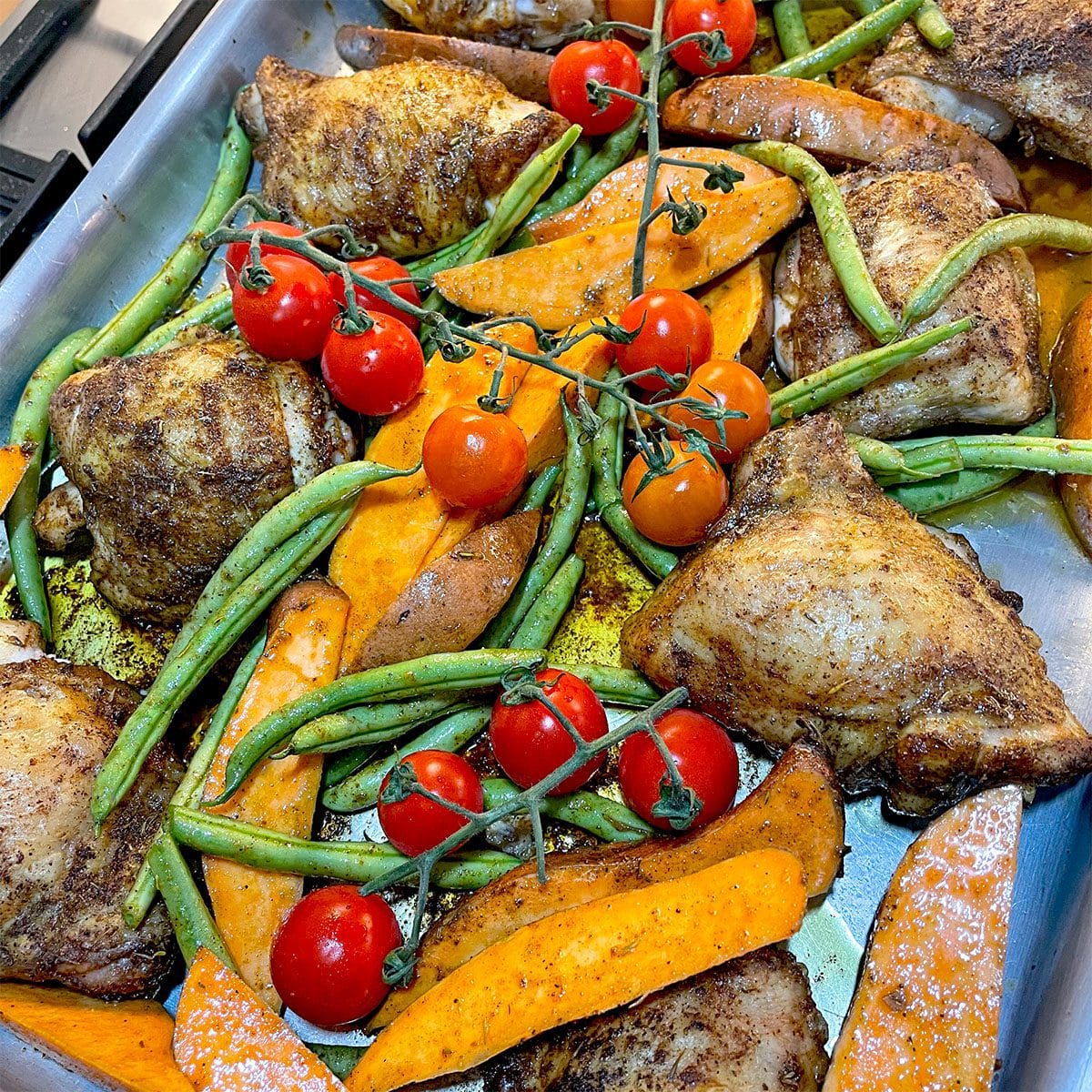 Baked chicken thighs, sweet potato wedges, green beans, and vine tomatoes arranged on a metal tray, seasoned and ready to eat.