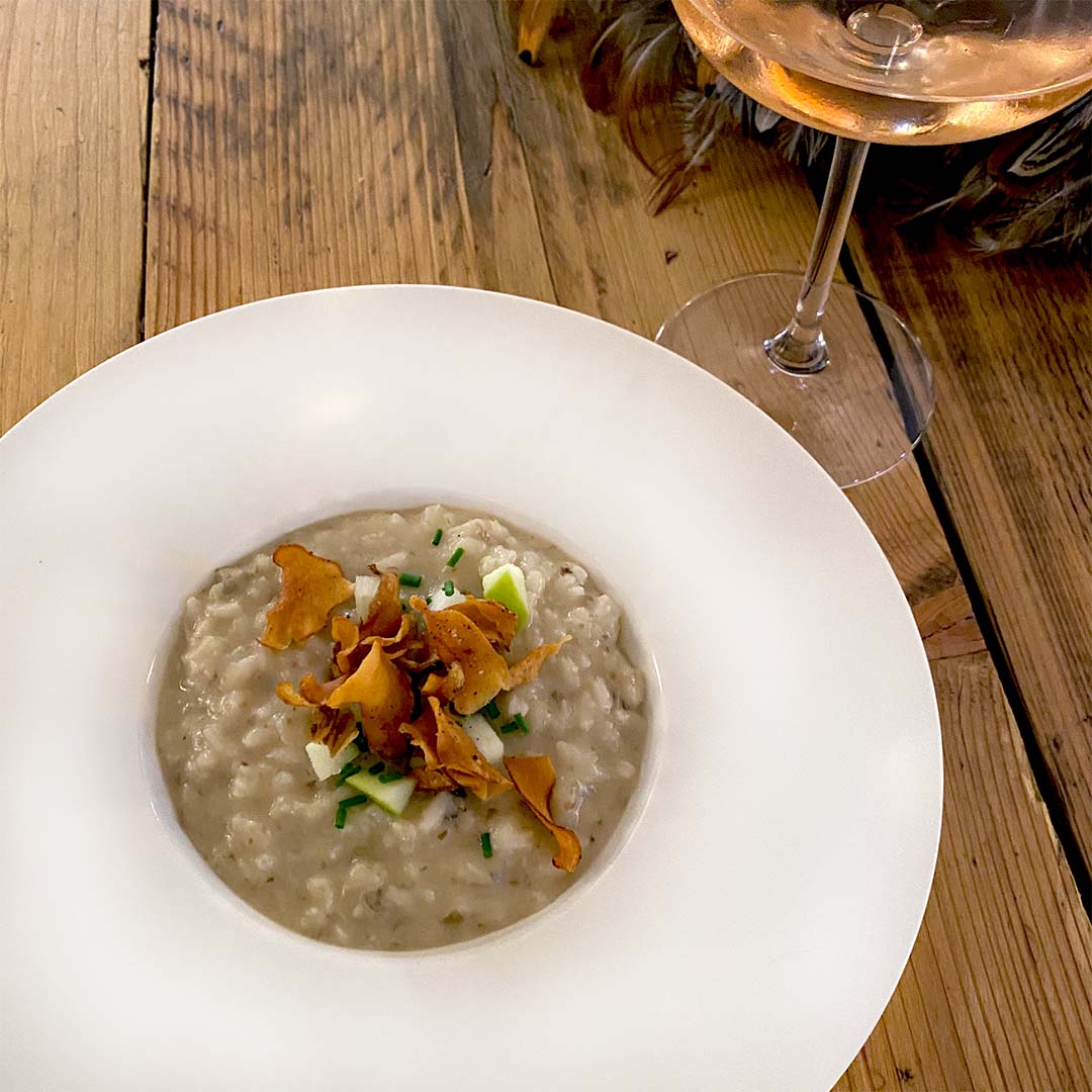 A white bowl of creamy risotto topped with crispy crisps and chopped herbs sits on a wooden table next to a glass of rosé wine.