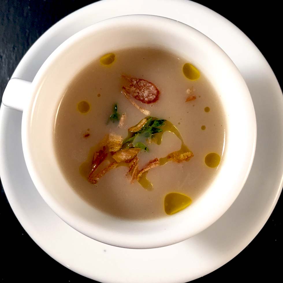 A white cup on a white saucer filled with creamy soup, garnished with crispy fried shallots, green herbs, and drops of oil, set on a dark surface.