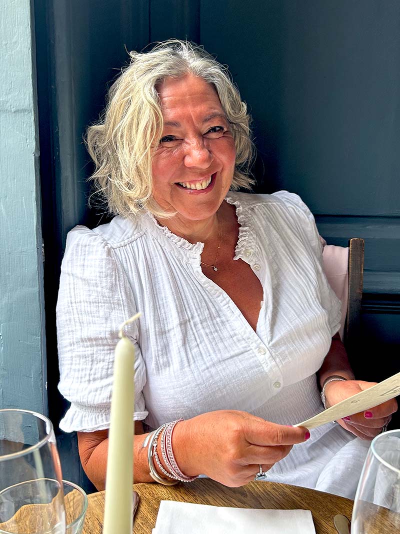 A smiling woman with wavy blonde hair wearing a white blouse sits at a table in a restaurant, holding a menu. A white candle and wine glasses are on the table in front of her.