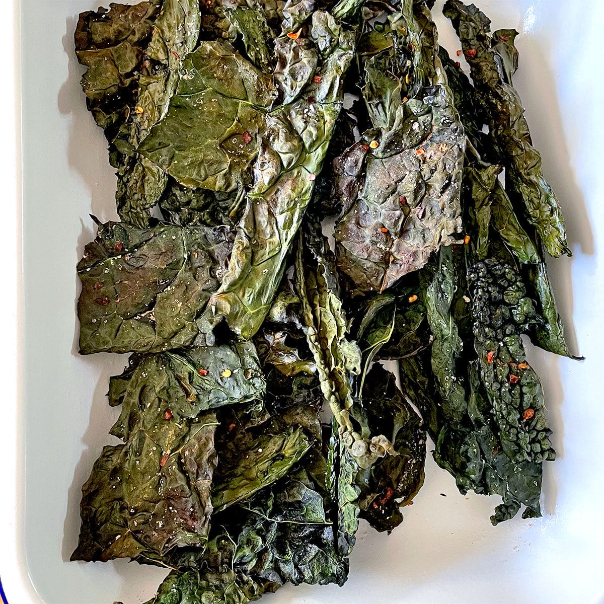 A white rectangular dish filled with crispy, baked kale crisps. The leafy crisps are dark green and slightly curled, with visible seasoning flakes scattered on their surfaces.