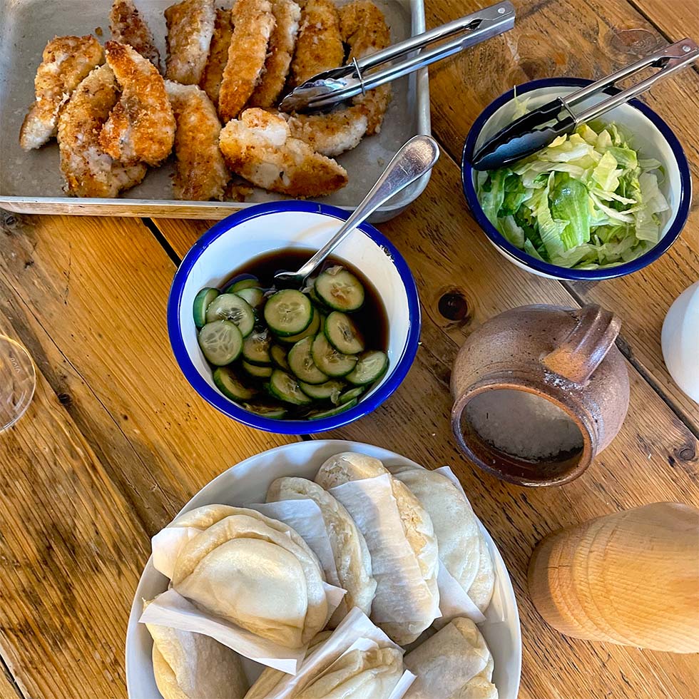 A wooden table set with a tray of breaded chicken goujons, a bowl of sliced cucumbers in sauce, a bowl of shredded lettuce, a pile of folded flatbreads, and a brown pottery jug.