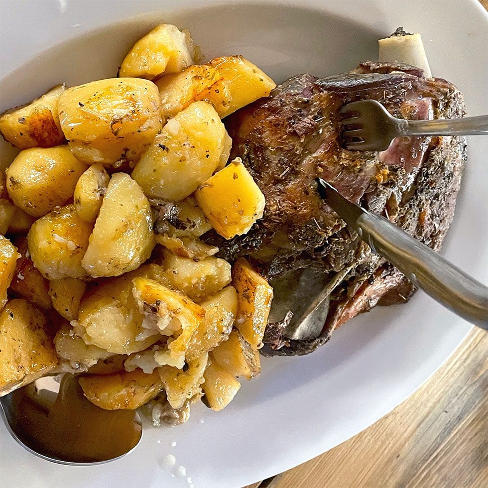 A white plate holding roast potatoes seasoned with herbs and a piece of roast meat with a fork and knife cutting into it, on a wooden table.
