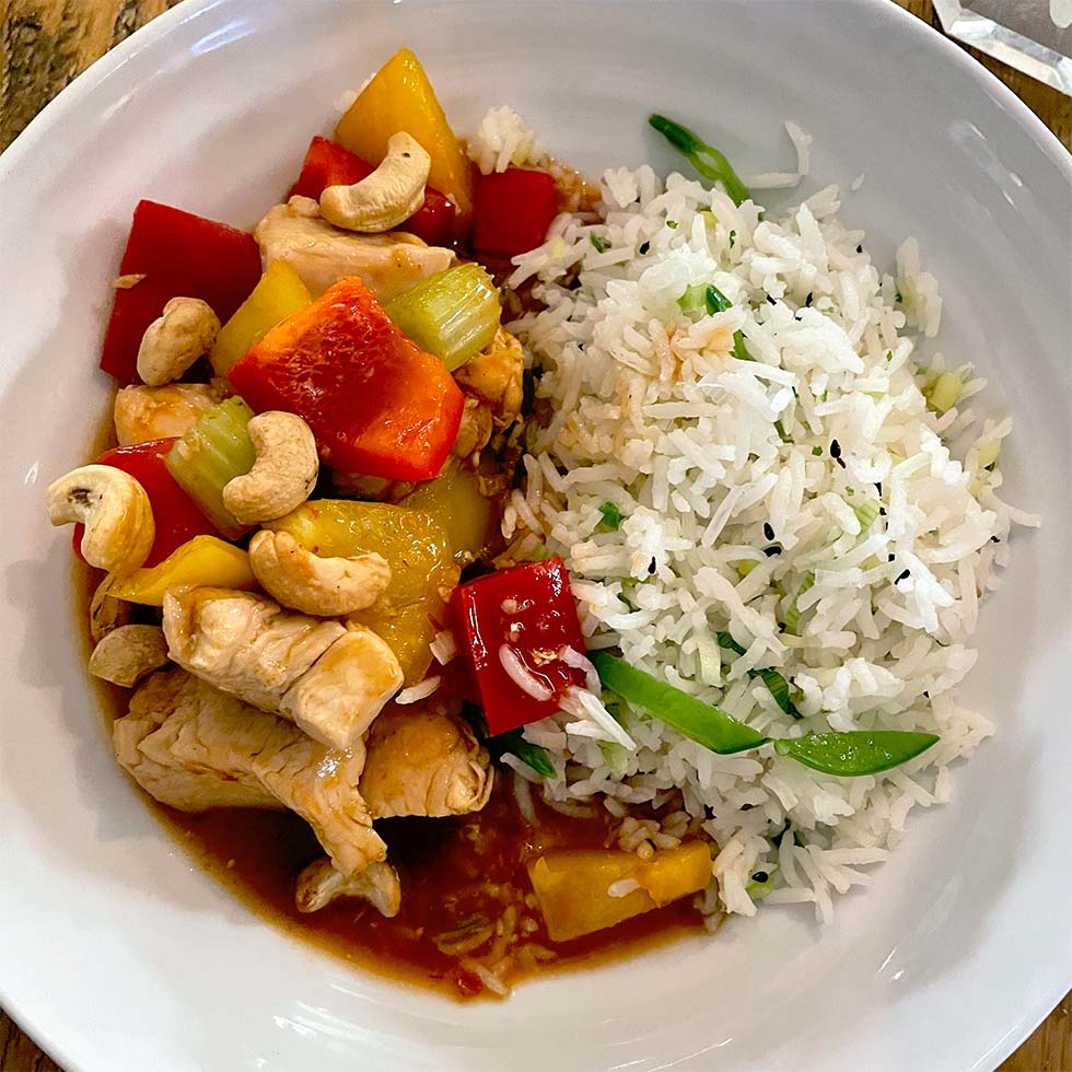 A white bowl filled with white rice mixed with vegetables, served alongside stir-fried chicken with red and yellow peppers, celery, pineapple, and topped with cashew nuts in a savoury sauce.
