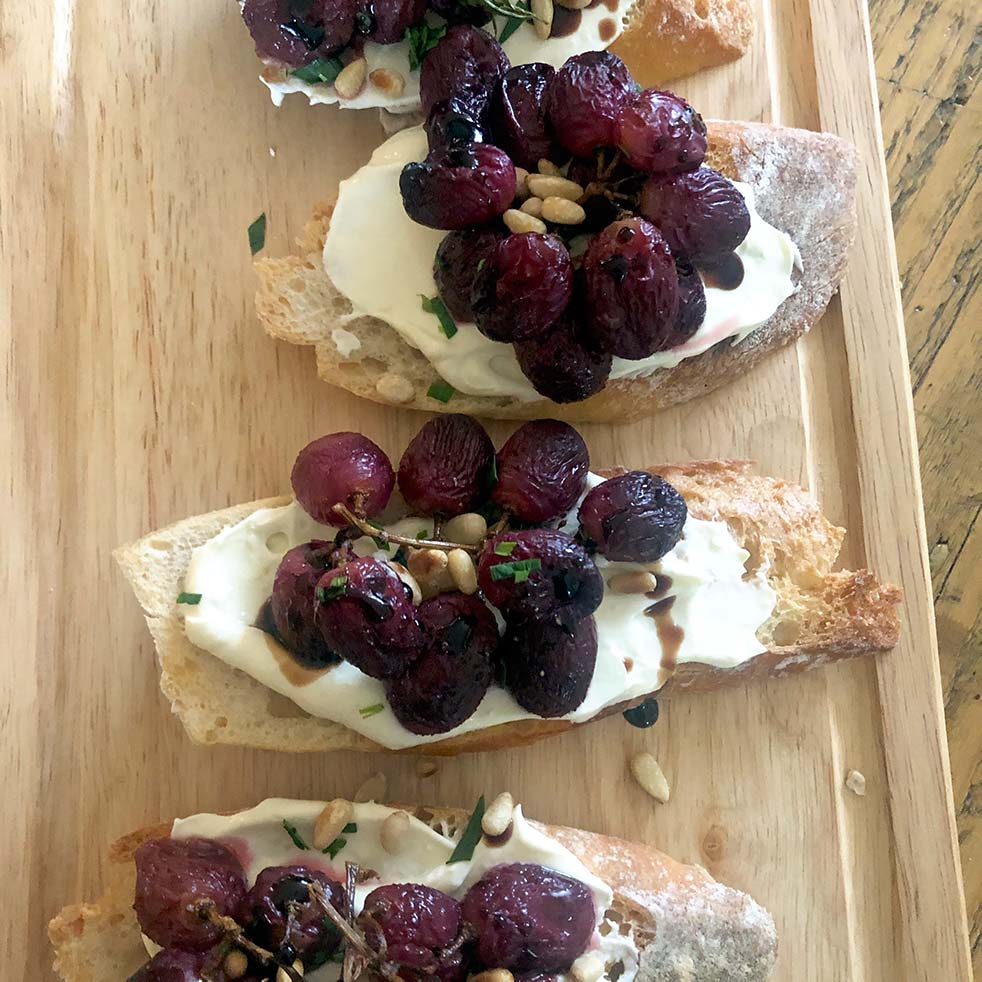 Three slices of crusty bread topped with creamy cheese, roasted grapes, pine nuts, and fresh herbs are arranged on a wooden serving board.