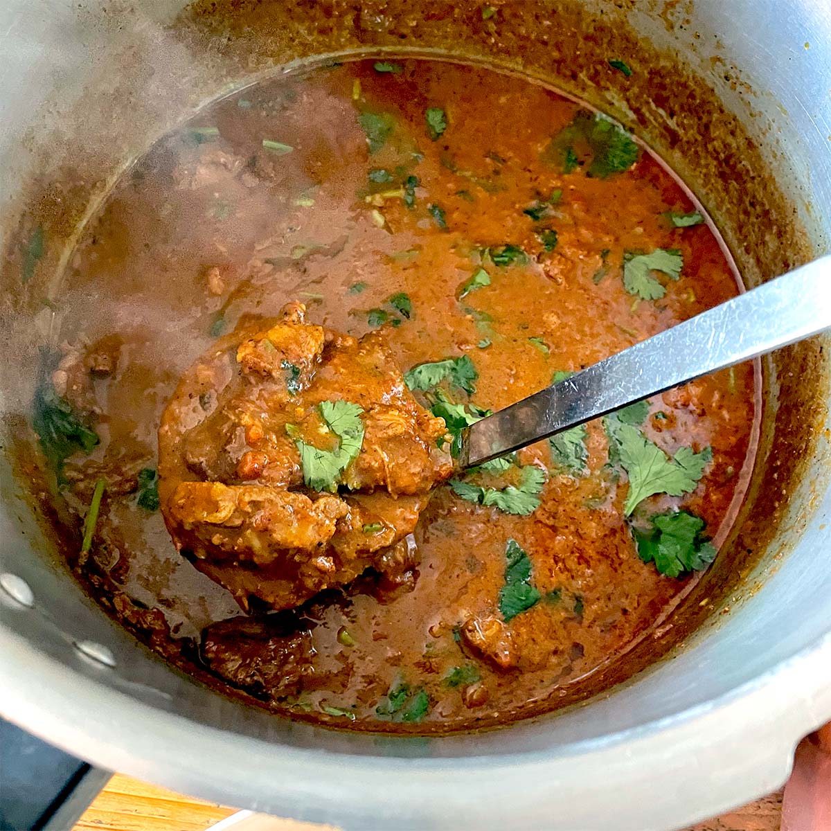 A pot of rich, red-brown curry garnished with fresh coriander, with a ladle lifting a portion of the meat and sauce. Steam rises from the hot dish, indicating it is freshly cooked.