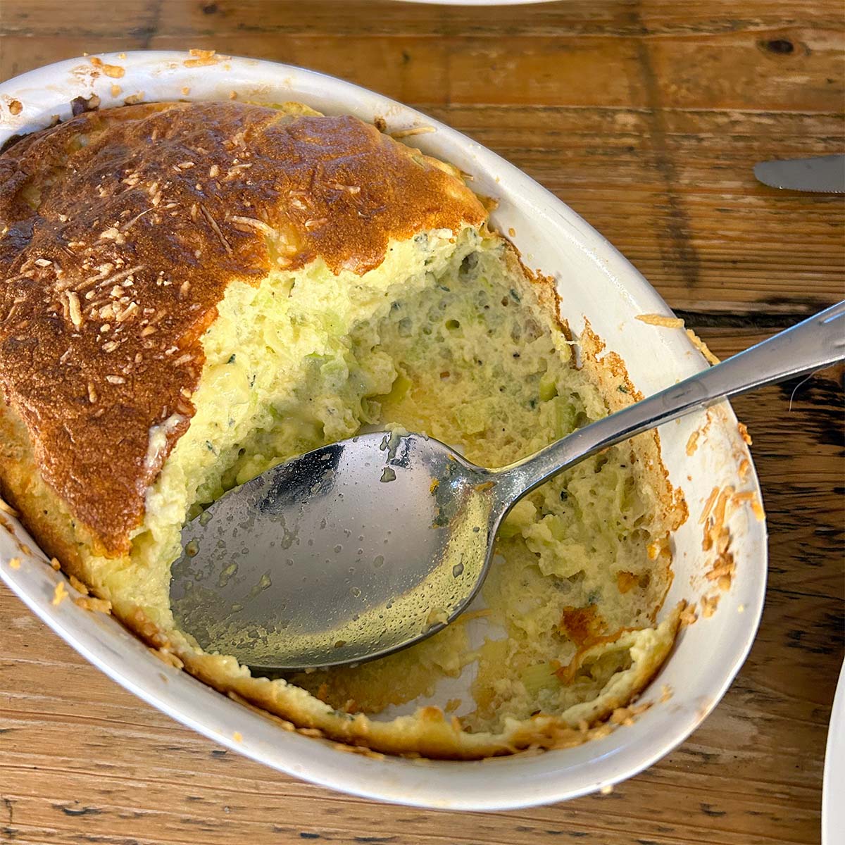 A close-up of a baked courgette soufflé in a white oval dish, with a spoon resting inside and part of the soufflé scooped out, showing its fluffy, airy texture. The dish sits on a rustic wooden table.