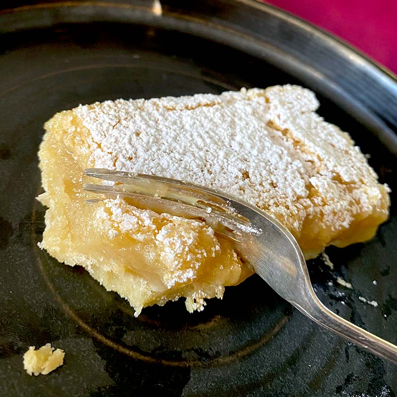 A slice of lemon bar covered with icing sugar sits on a dark plate, with a fork cutting into its soft, creamy texture.