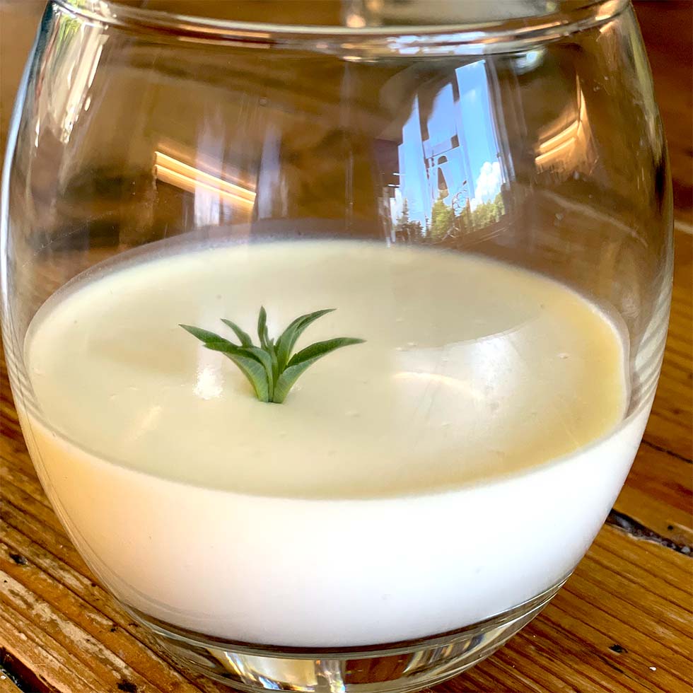 A round glass filled with a creamy white dessert, topped with a small green herb sprig, sits on a wooden table. The background is softly reflected on the glass.