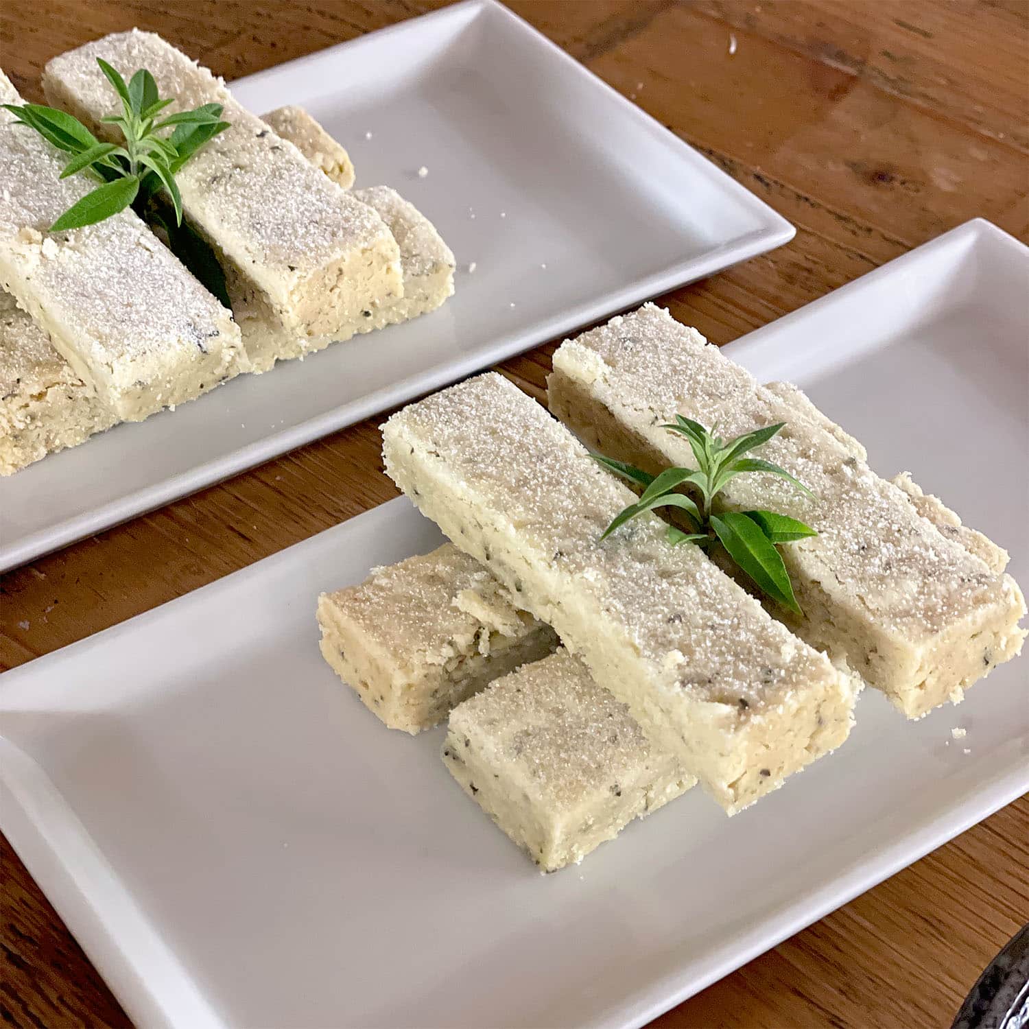Rectangular pieces of shortbread are neatly stacked on two white rectangular plates, dusted with icing sugar and garnished with a small sprig of green herb. The plates rest on a wooden surface.
