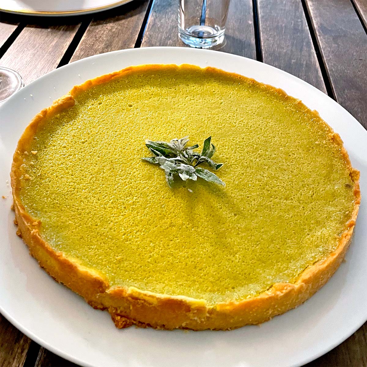 A round matcha green tea tart with a golden crust, topped with a small cluster of sugared mint leaves, sits on a white plate on a wooden table.