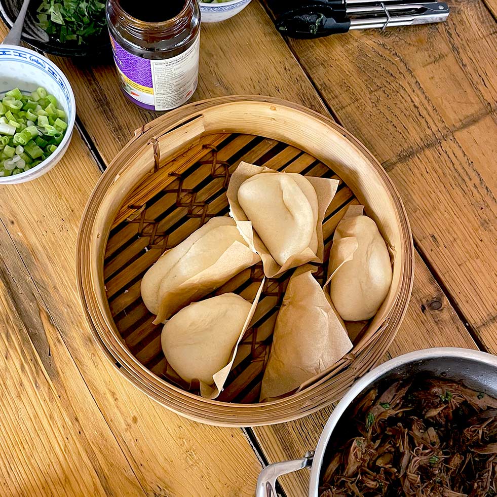 A bamboo steamer with five folded bao buns sits on a wooden table, surrounded by bowls of spring onions, herbs, a jar of sauce, tongs, and a pot of shredded meat.