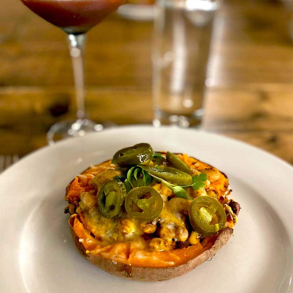 A baked sweet potato half topped with melted cheese, beans, and sliced jalapeños sits on a white plate. In the background are a glass of red drink and a tall glass of water on a wooden table.