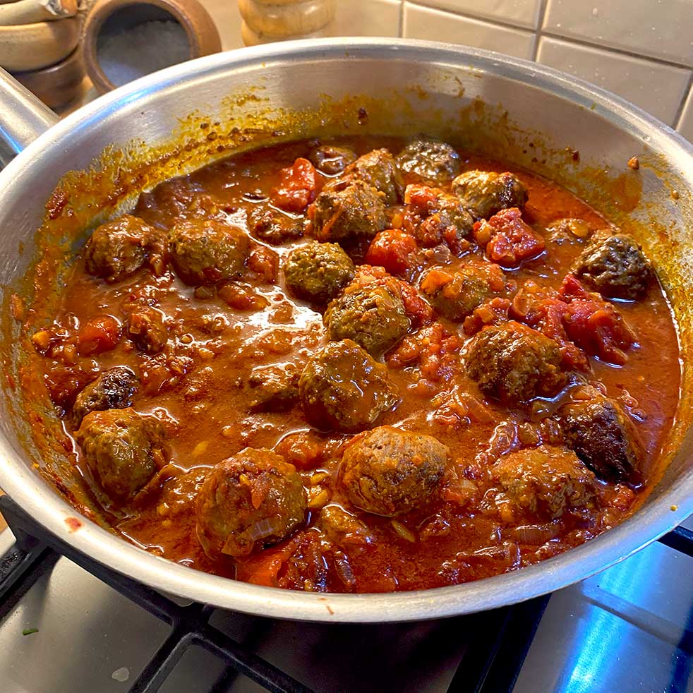 A frying pan filled with meatballs simmering in a rich tomato sauce with chunks of tomatoes, set on a hob with tiled splashback in the background.