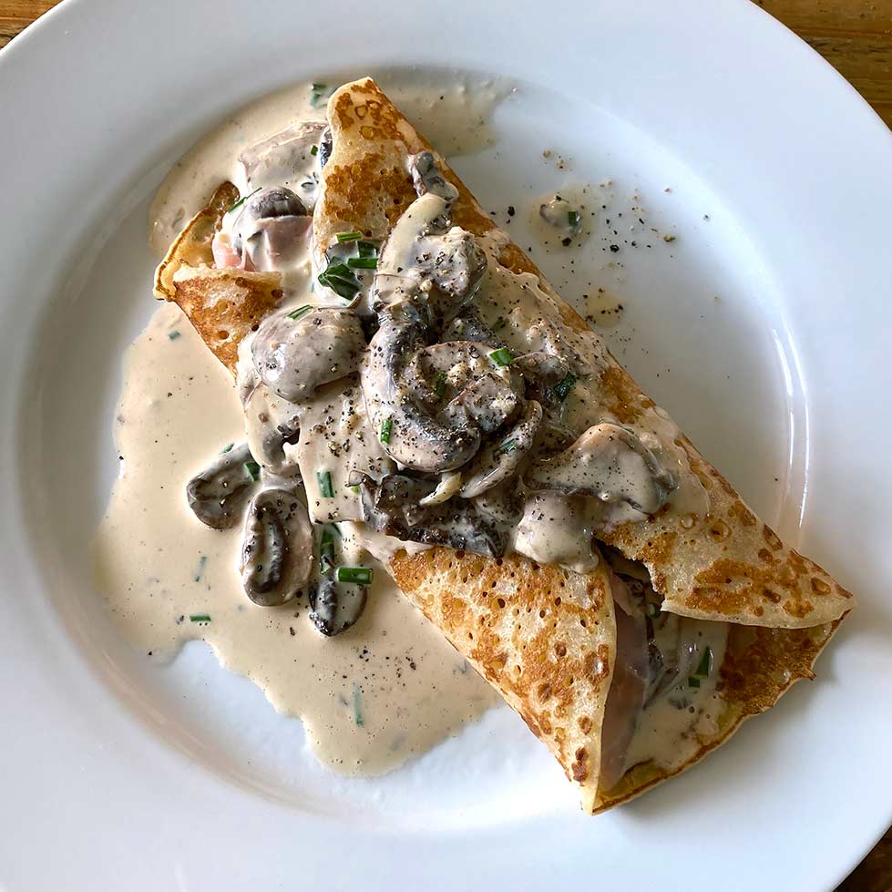 A savoury crêpe filled with mushrooms and topped with creamy mushroom sauce and chopped herbs, served on a white plate.