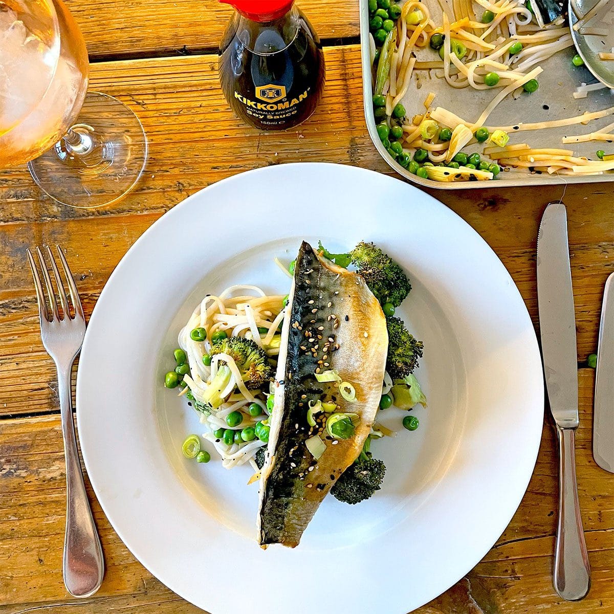 A white plate with grilled fish on top of noodles, broccoli, peas, and bean sprouts. A fork and knife are on either side, with a glass of drink and a bottle of soy sauce on a wooden table.