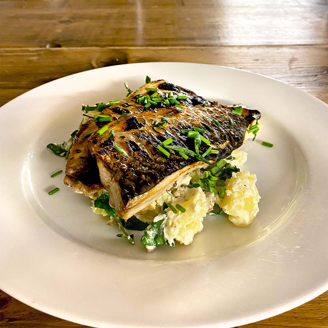 Grilled fish fillet garnished with chopped chives, served on a bed of salad leaves and creamy potato salad, presented on a white plate atop a wooden table.