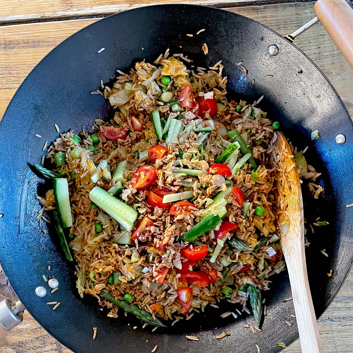 A wok filled with fried rice mixed with peas, tomatoes, cucumber slices, greens, and minced meat, with a wooden spoon resting on the side. The dish sits on a wooden table.