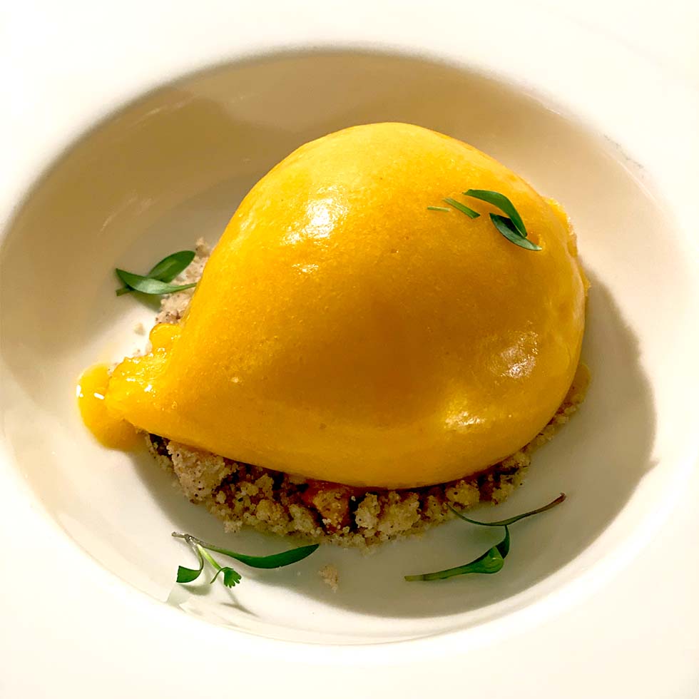 A bright yellow scoop of mango sorbet sits on a bed of crumbled biscuit, garnished with a few small green herb leaves, served in a white bowl.