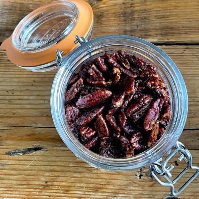 A glass jar filled with roasted pecan halves sits open on a rustic wooden surface, with the orange-rimmed lid resting to the side and attached by a metal clasp.