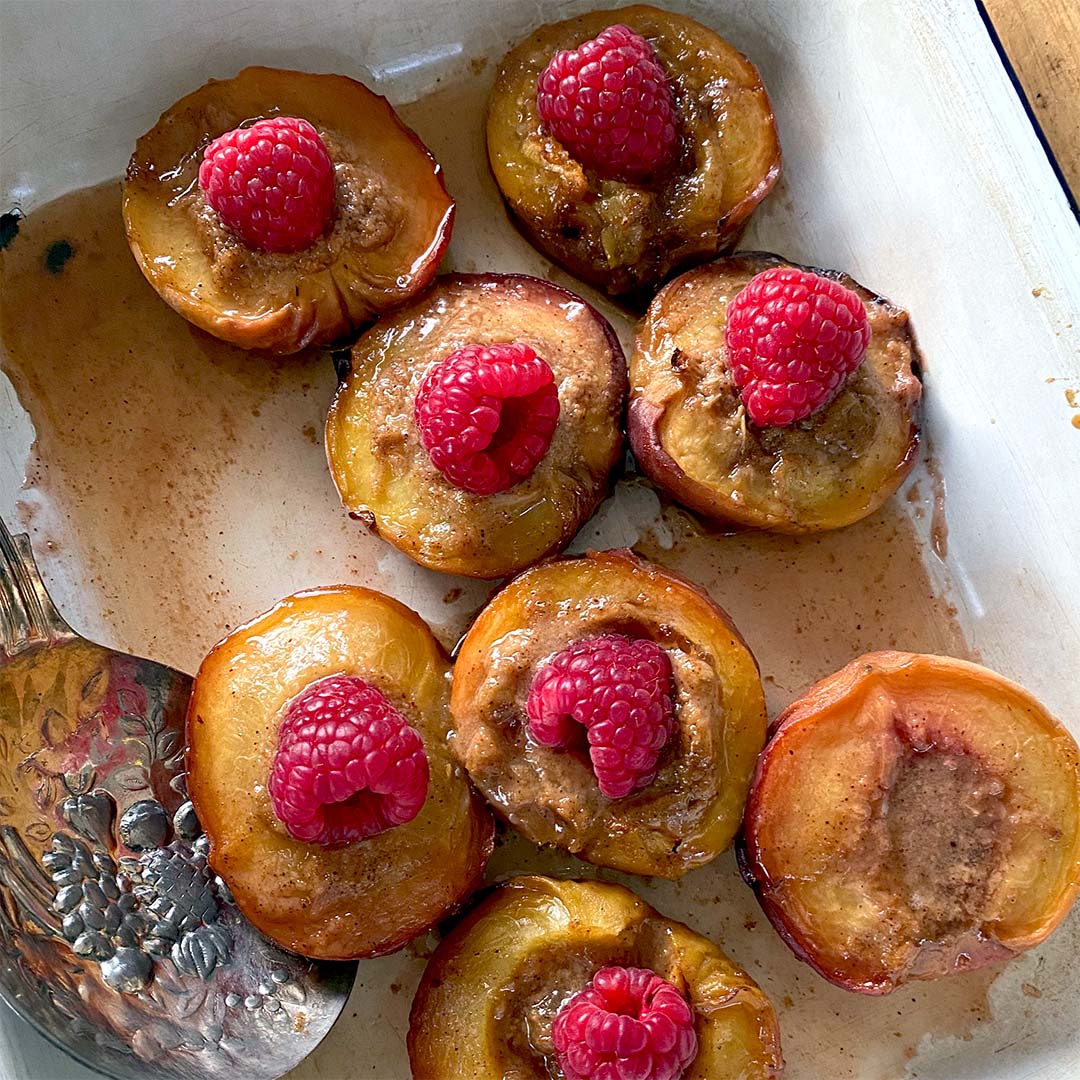 Halved baked peaches topped with a dollop of brown filling and fresh raspberries, arranged in a white baking dish with a serving spoon at the side.