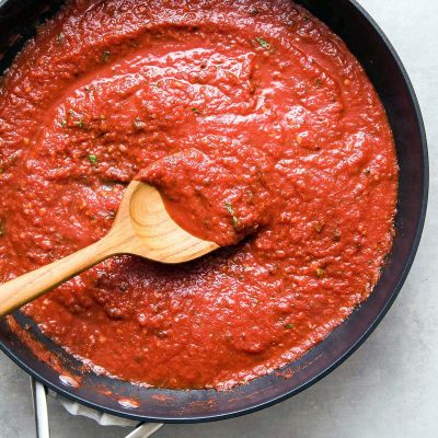 A wooden spoon stirs a thick, red tomato sauce with herbs in a black pan, viewed from above.