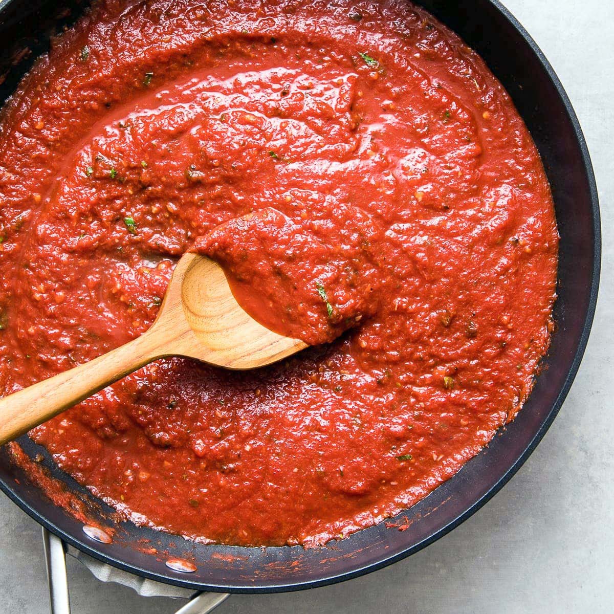 A wooden spoon stirs a thick, red tomato sauce with herbs in a black pan, viewed from above.